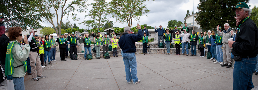 A large group of Lamorinda CERT volunteers in green safety vests gathered in a circle outdoors during a community disaster-response briefing.